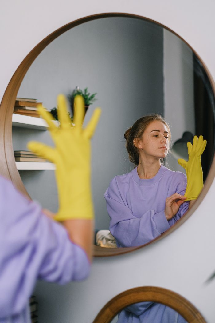 A woman wearing yellow gloves is reflected in a wall mirror indoors, exuding a serene expression.